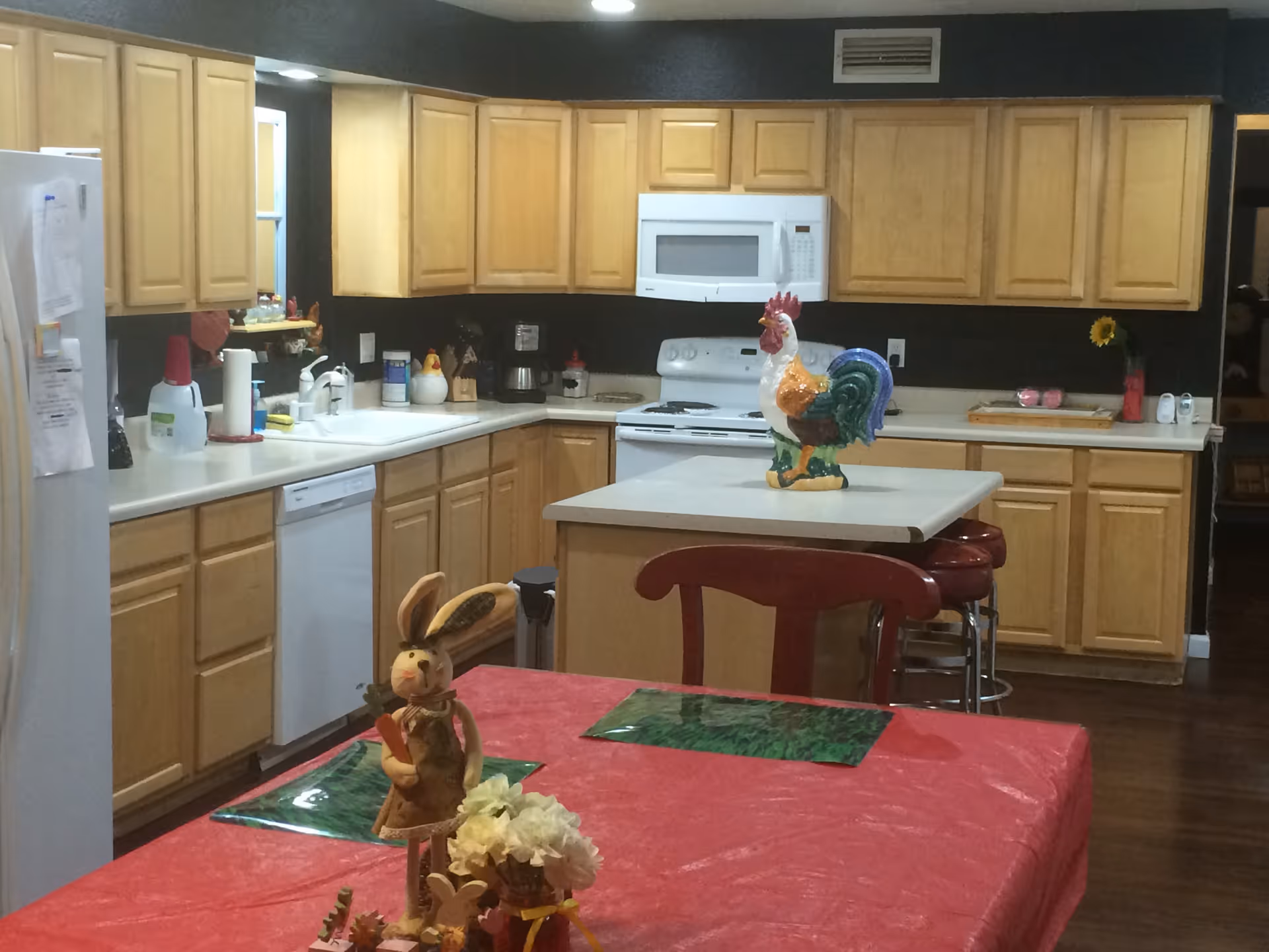 A kitchen with light wood cabinets, white countertops, and white appliances including a refrigerator, dishwasher, stove, and microwave. A kitchen island with a colorful rooster decoration is in the center. In the foreground, there is a table covered with a red tablecloth, decorated with a bunny figurine and a small flower arrangement. The floor is dark wood, and the walls are painted dark.
