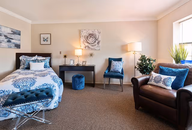 A cozy senior living bedroom featuring a single bed with blue and white patterned bedding, a dark wooden desk with decorative items and a lamp, a blue upholstered chair with a patterned pillow, a brown leather armchair with blue and white pillows, a small blue ottoman, and a window letting in natural light. The walls are decorated with framed artwork including a butterfly and a large flower.