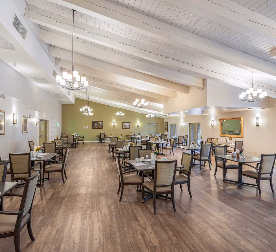 Spacious dining room with wooden floors, multiple tables and chairs arranged neatly, decorated with small flower vases and place settings. The room features high white beamed ceilings with modern chandeliers, light-colored walls with framed artwork, and a large mirror on one side.