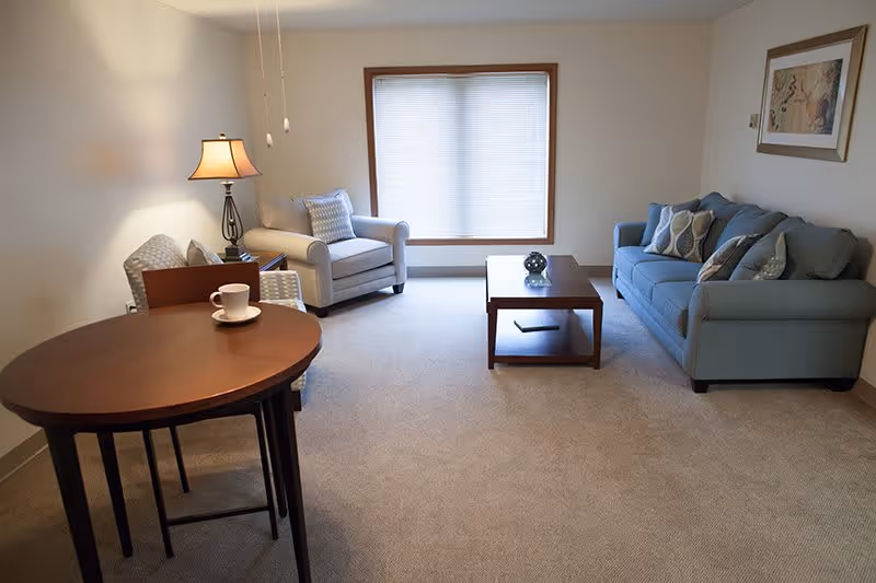 A cozy living room with a blue sofa adorned with patterned pillows, a beige armchair, a wooden coffee table with a decorative item, a round wooden dining table with a cup and saucer, a floor lamp, and a large window with closed blinds.