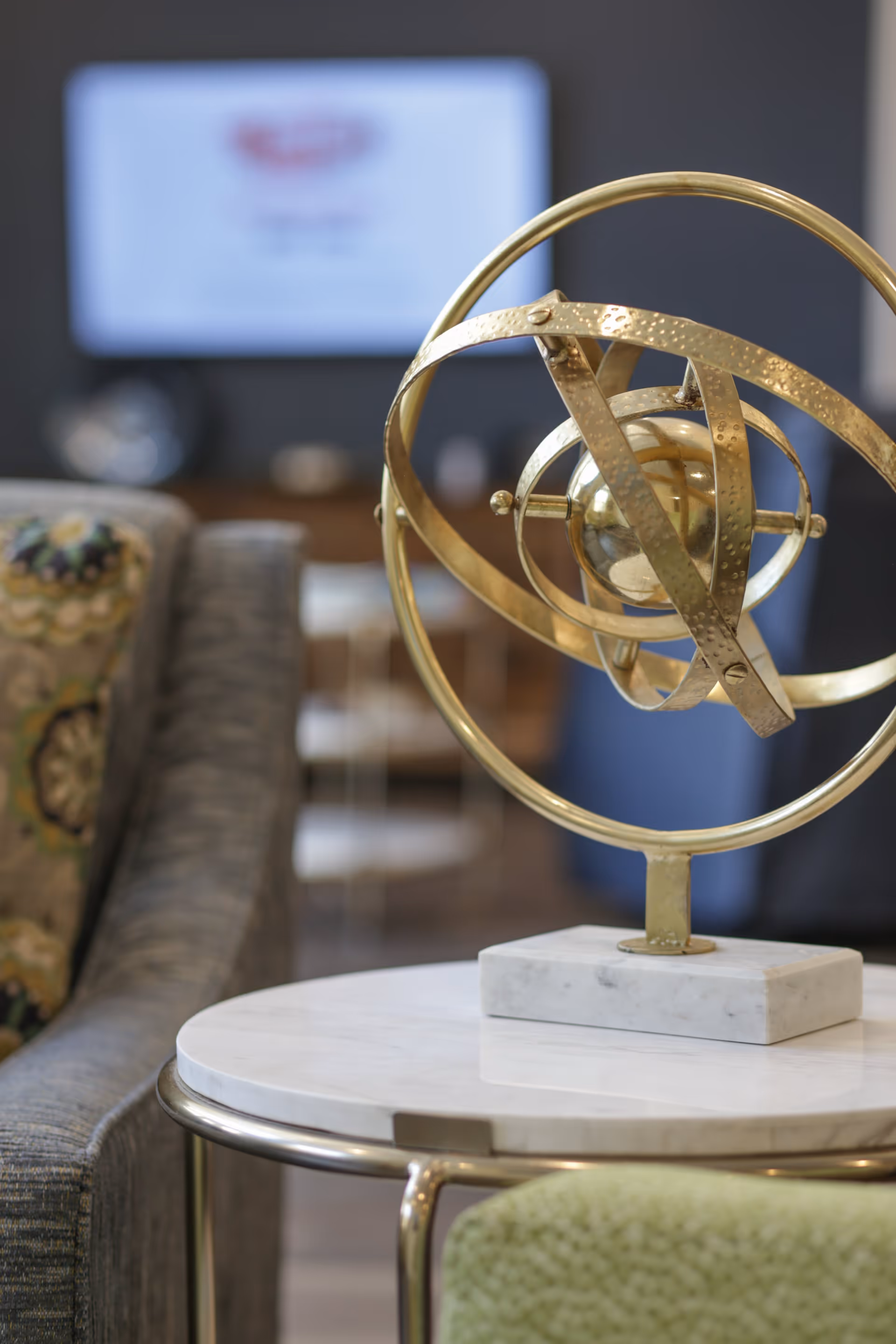Close-up of a decorative golden armillary sphere on a white marble table in a living room setting with a patterned cushion on a gray sofa and a blurred TV screen in the background.