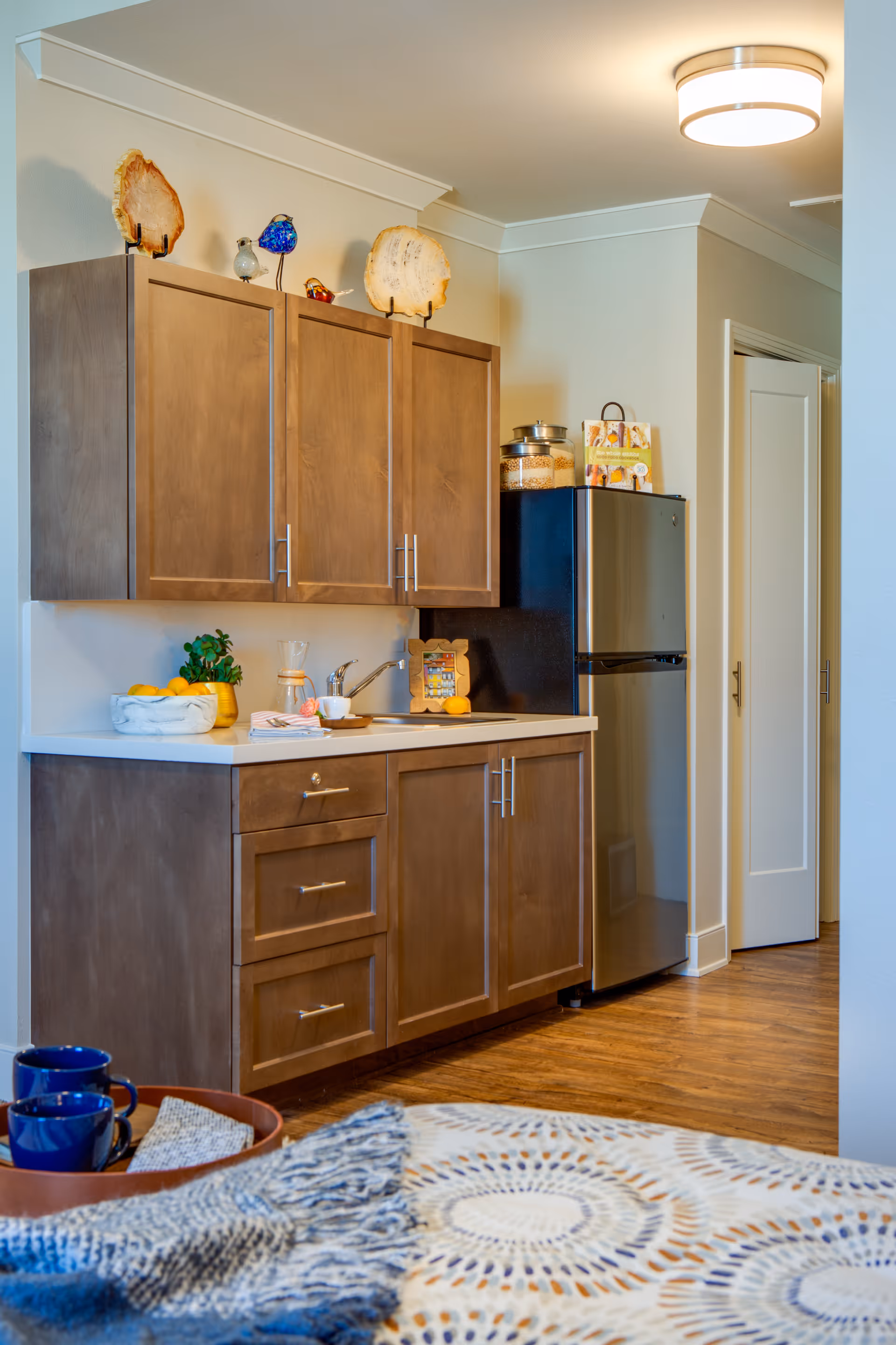 A small kitchen area with wooden cabinets, a countertop with a sink, a stainless steel refrigerator, and decorative items on top of the cabinets. The floor is wooden, and part of a bed with a patterned blanket and a blue throw is visible in the foreground.