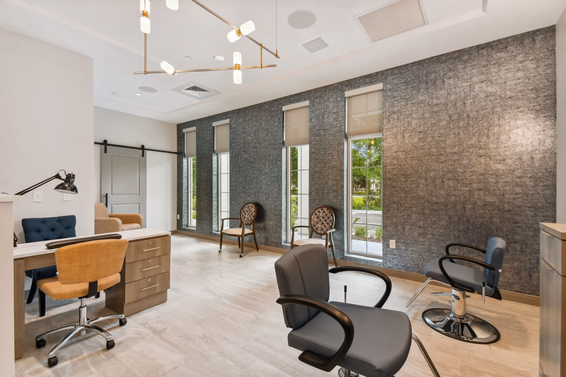 A modern salon area in a senior living facility with two gray salon chairs, a wooden desk with a rolling chair, two decorative chairs against a textured gray wall, and tall windows with beige roller shades letting in natural light.