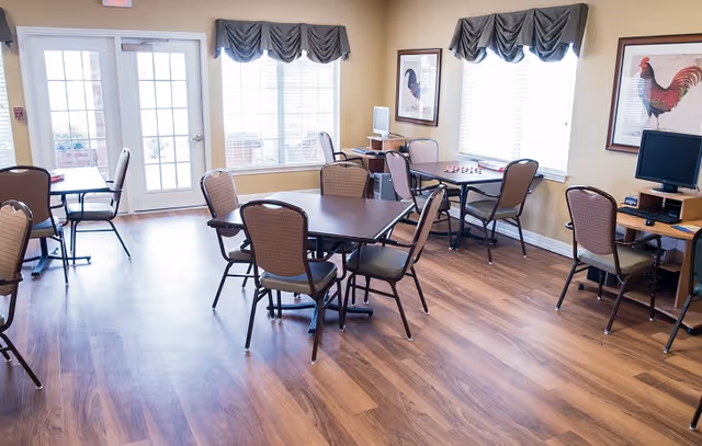 A bright room with several tables and chairs arranged for group seating, two computer desks with monitors, large windows with valance curtains, and framed rooster artwork on the walls.