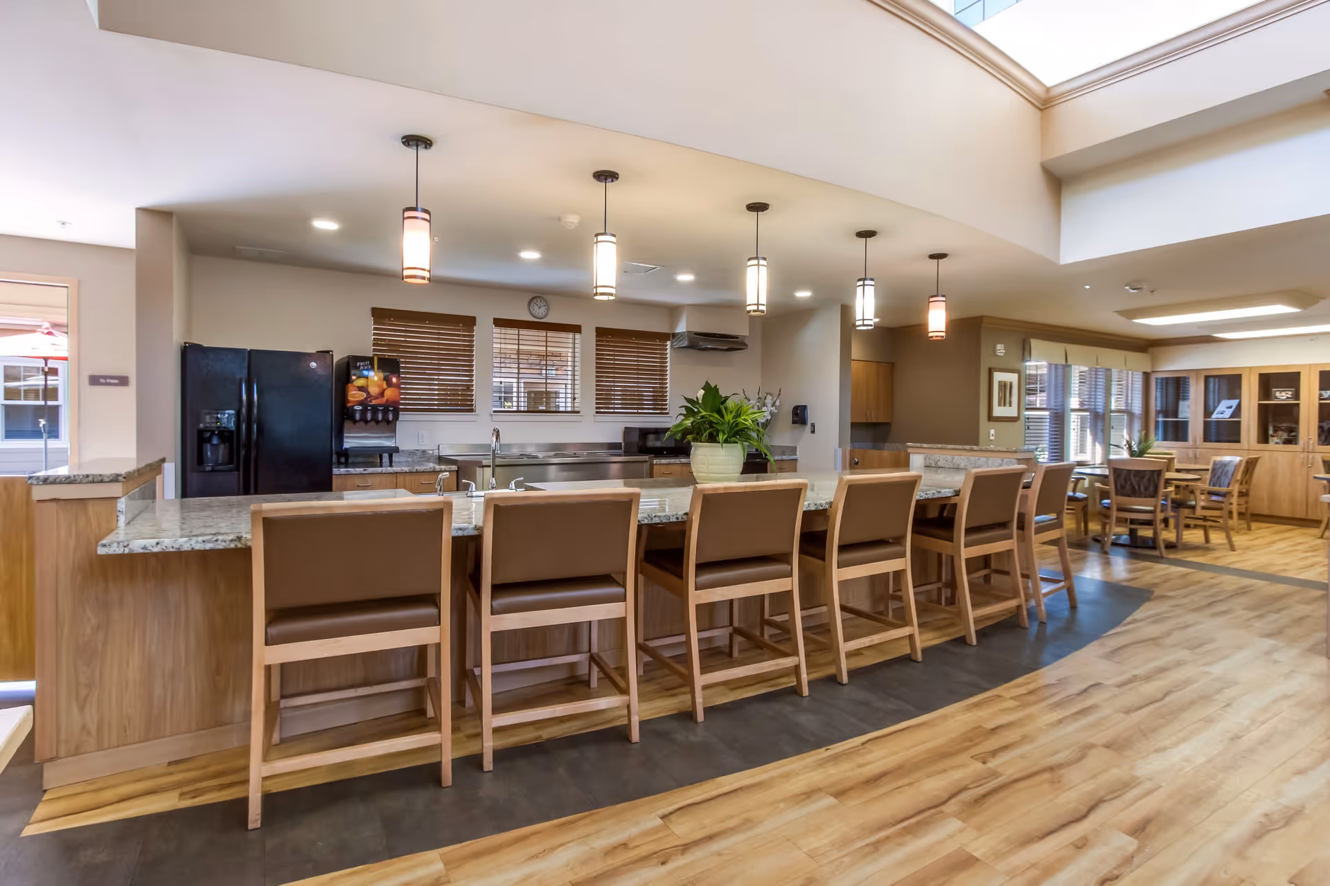 A spacious kitchen area in a senior living facility featuring a long granite countertop with seven wooden chairs. The kitchen includes a black refrigerator, a beverage dispenser, a sink, and several hanging pendant lights. In the background, there is a dining area with tables and chairs, wooden cabinetry, and large windows with blinds allowing natural light to enter.