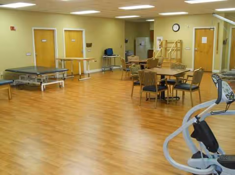 An activity room in a senior facility with exercise equipment, tables, and wooden flooring.