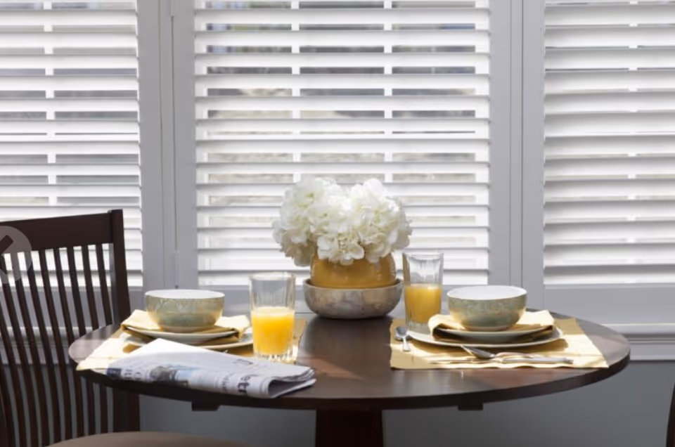 A small round wooden dining table set for two with two bowls on plates, two glasses of orange juice, silverware, and a folded newspaper. A yellow vase with white flowers is in the center of the table. The background shows white window shutters.