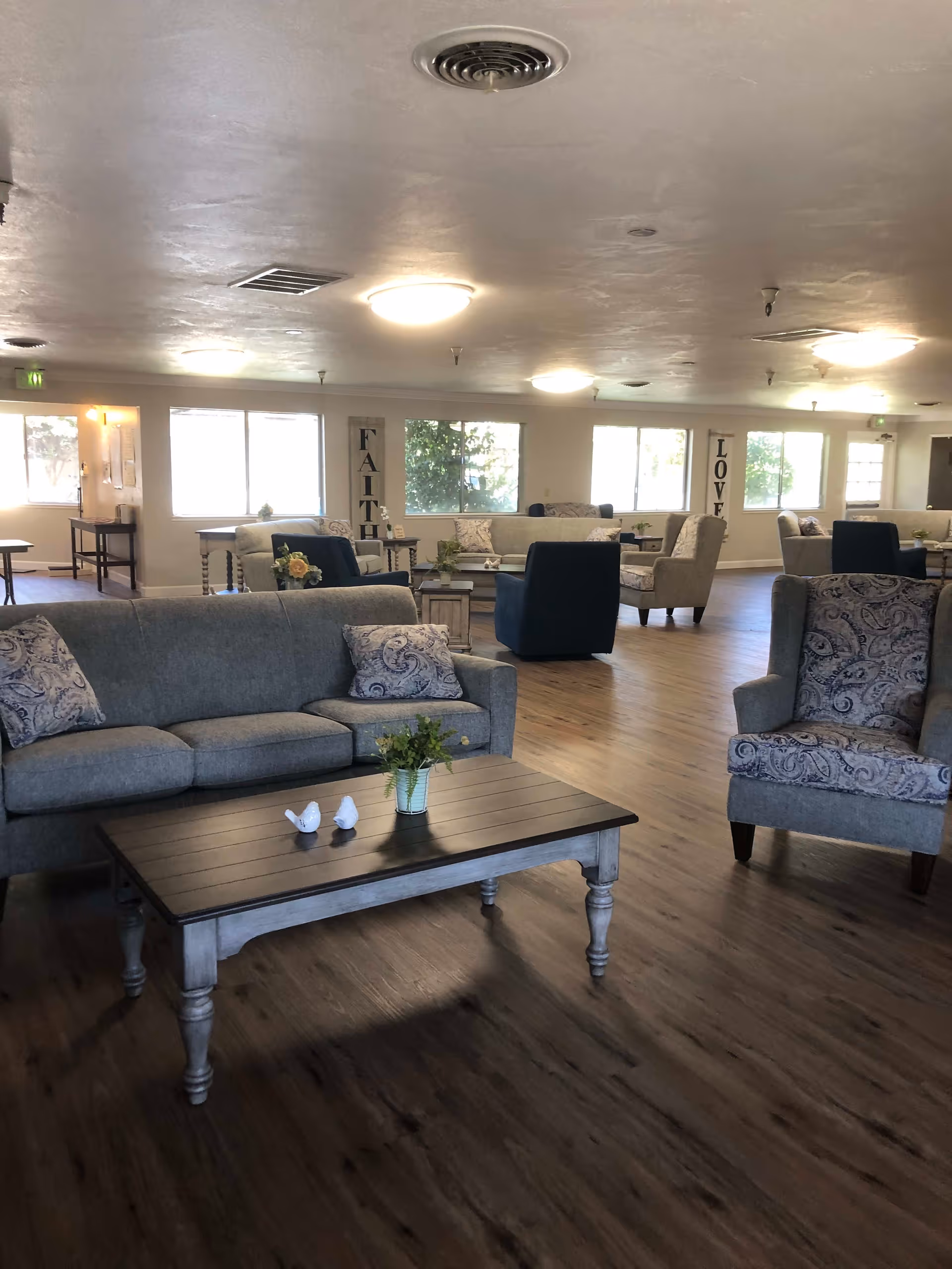 A spacious living room area in an assisted living facility with multiple sofas and armchairs arranged around wooden coffee tables. The room has large windows letting in natural light, wooden flooring, and decorative signs on the walls that say 'FAITH' and 'LOVE'.
