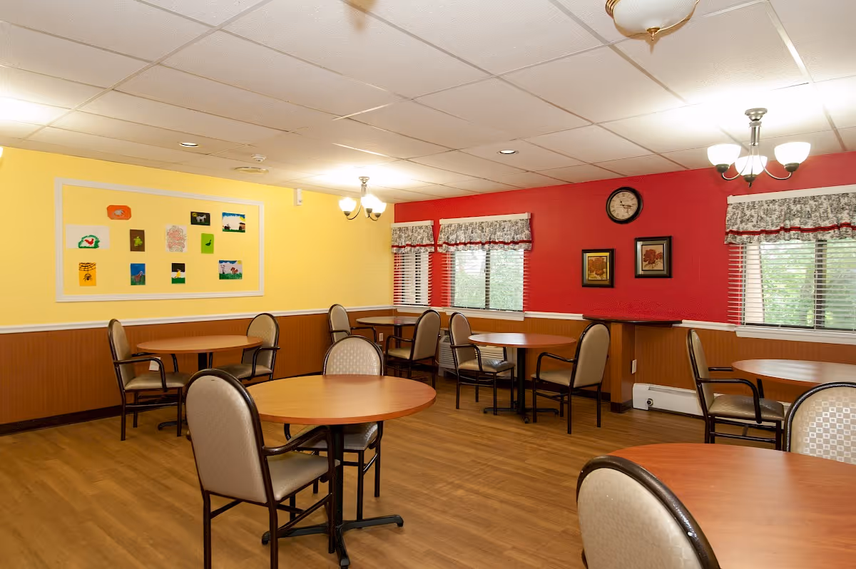A dining room with several round wooden tables and cushioned chairs arranged around them. The walls are painted yellow and red, with framed artwork and a clock on the red wall. There are windows with floral valances and blinds, and ceiling lights providing illumination.