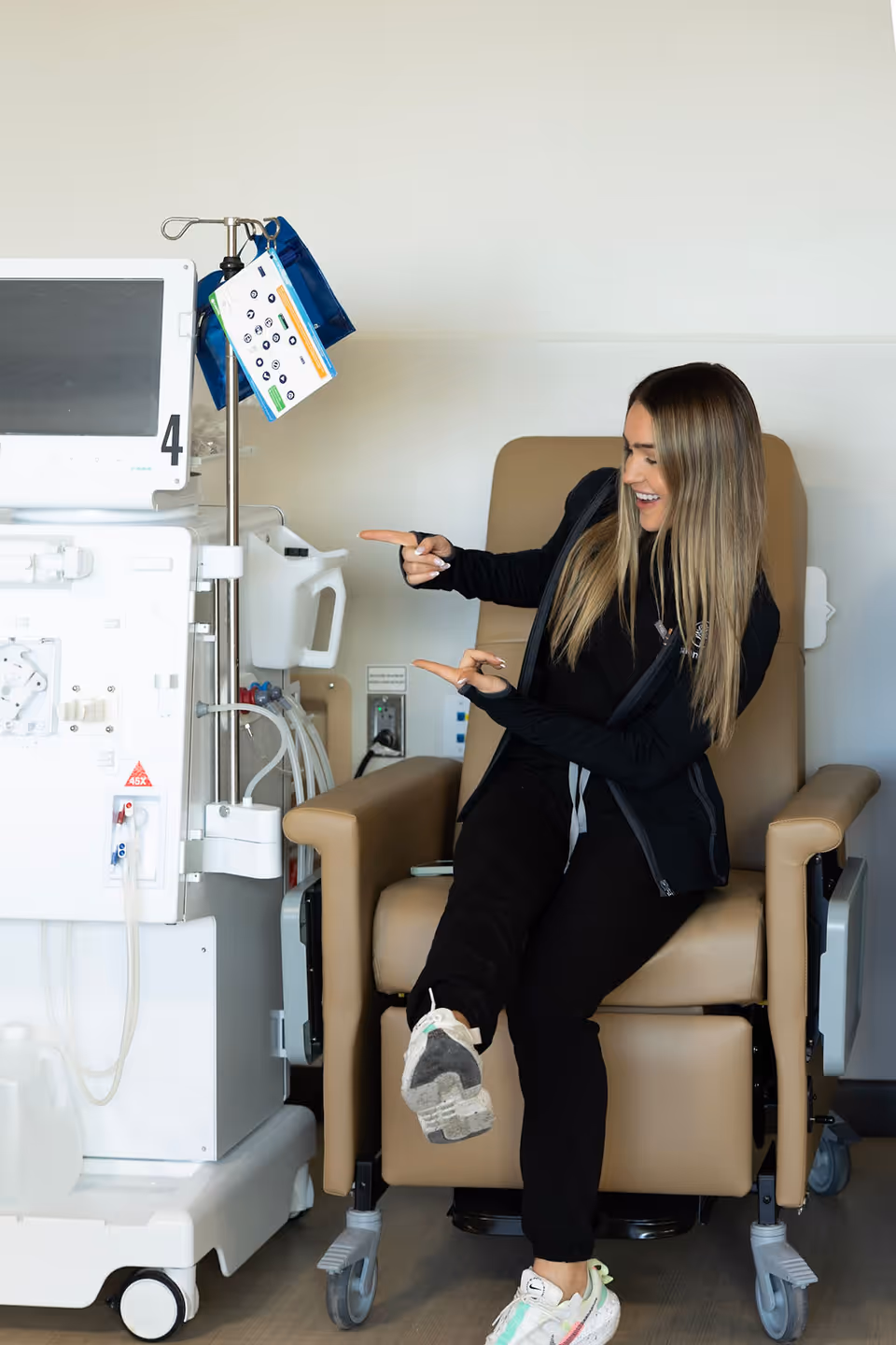 A woman with long blonde hair sitting in a beige medical recliner chair, smiling and pointing with both hands towards a dialysis machine next to her in a clinical setting.