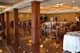 Spacious dining room with polished wooden floors, multiple wooden tables and chairs arranged neatly, decorative wooden columns, warm lighting fixtures on the ceiling, and a buffet table covered with a white tablecloth on the right side.