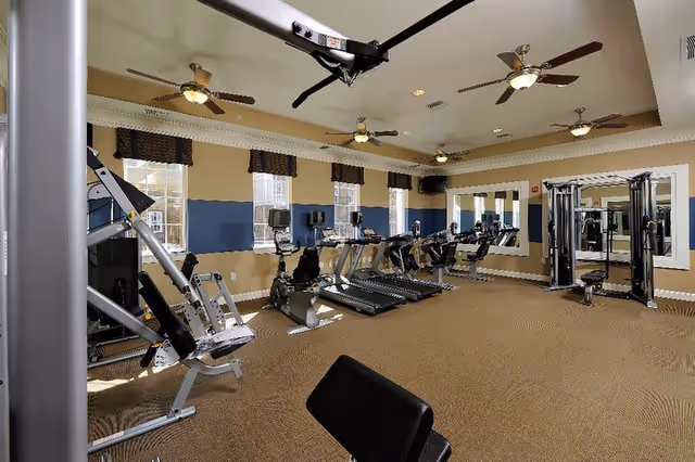A spacious fitness room with various exercise equipment including treadmills, stationary bikes, and weight machines. The room has beige walls with a blue accent stripe, multiple windows with valances, ceiling fans, and large mirrors on one wall.