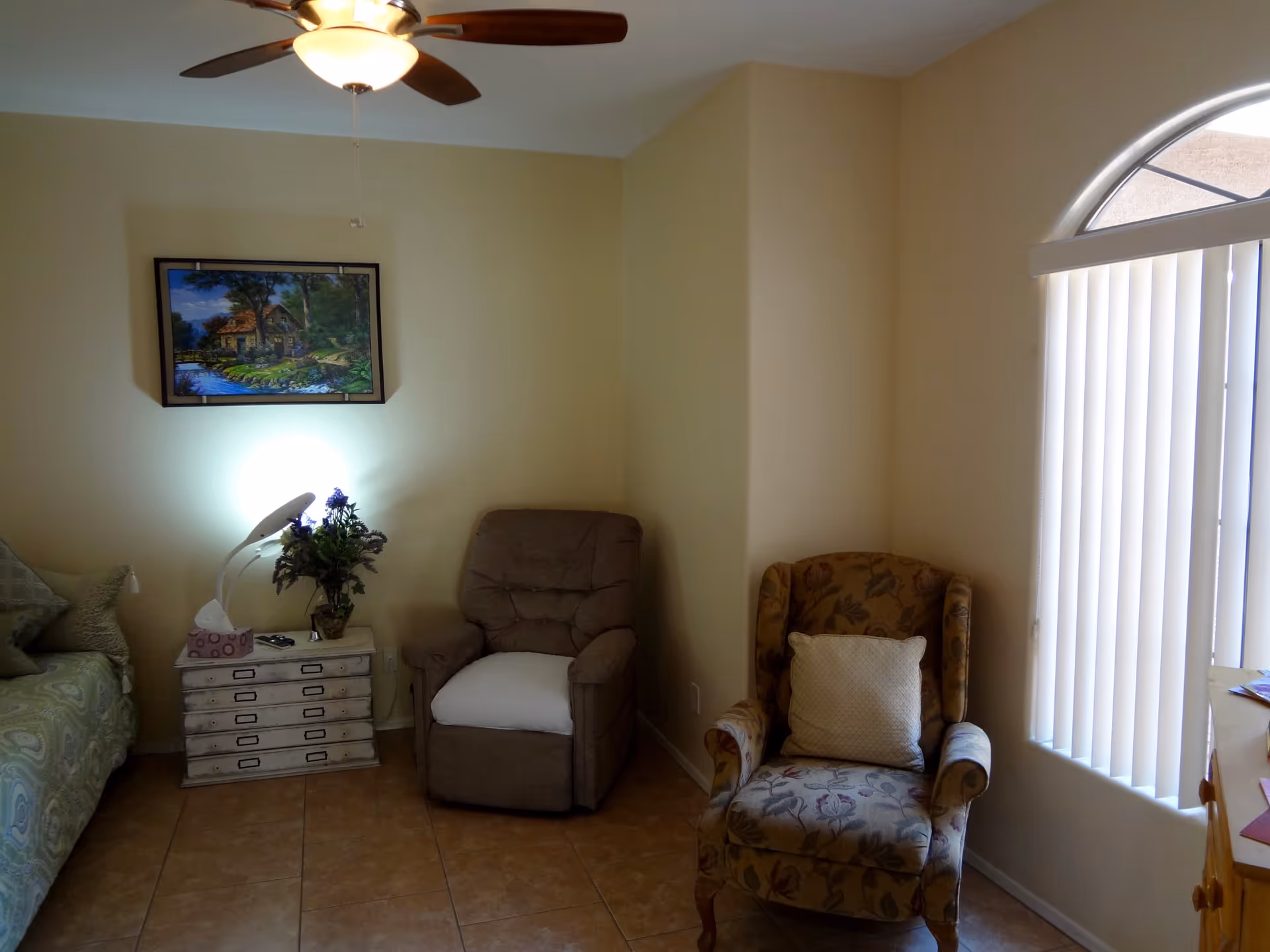 A cozy corner of a living room with two armchairs, one brown recliner and one floral patterned chair with a white cushion. There is a small white side table between the chairs with a lamp, a vase of flowers, and a tissue box on it. A framed painting of a house by a river hangs on the beige wall above the side table. A ceiling fan with a light is visible on the ceiling, and a window with vertical blinds lets in natural light.