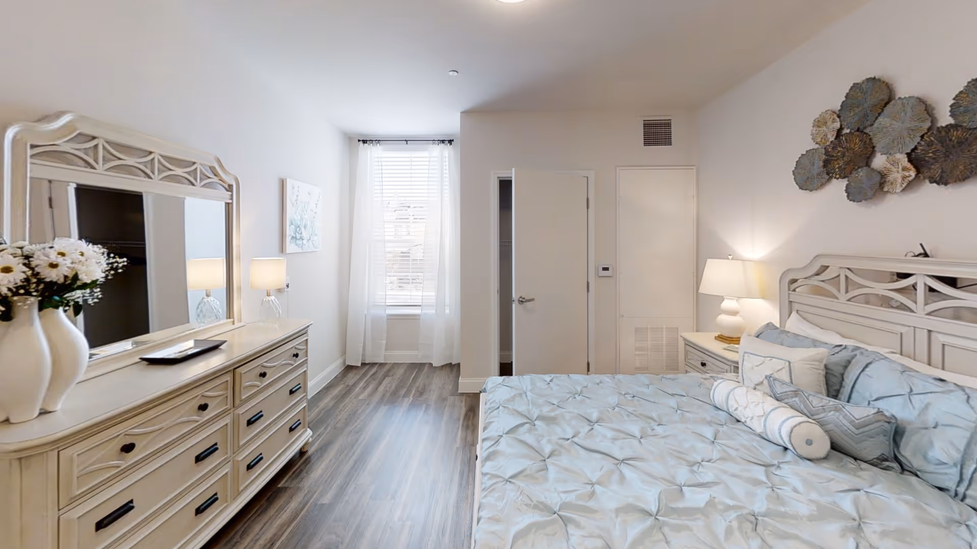 A bright and airy bedroom featuring a large bed with light blue bedding and multiple pillows. The room has a wooden dresser with a large mirror, a vase with white flowers, and two table lamps on either side of the bed. There is a window with sheer white curtains allowing natural light to fill the space. The walls are white, and the floor is a dark wood laminate. Decorative wall art is hung above the bed.
