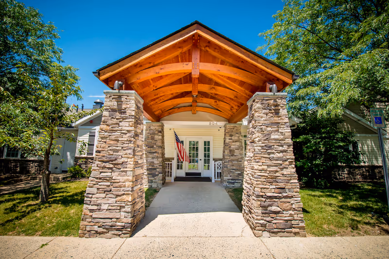 Entrance of a building with a wooden peaked roof supported by stone pillars, an American flag hanging near the white double doors, and greenery on both sides under a clear blue sky.