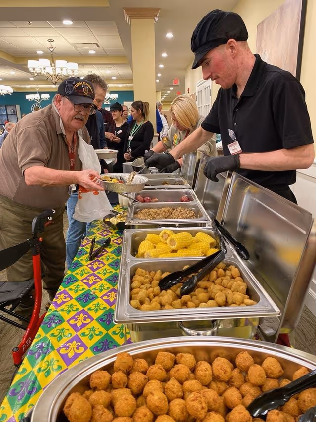 People at a buffet table in a dining room serving and receiving trays of food including hush puppies and corn on the cob.