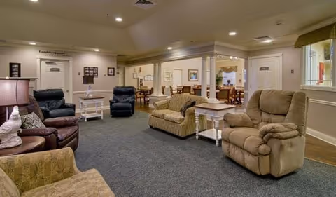 A spacious senior living common area with multiple comfortable armchairs and sofas arranged around small side tables. The room has carpeted flooring, soft lighting, and a dining area visible in the background with wooden tables and chairs. The walls are light-colored with framed artwork and decorative molding.