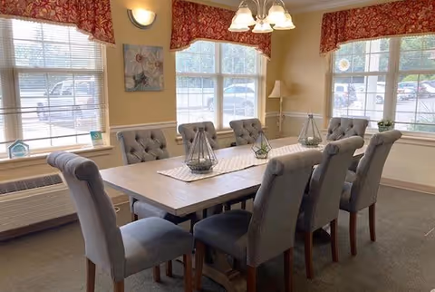 Sunlit dining room with a rectangular wooden table surrounded by eight tufted gray upholstered chairs and windows with red valances.