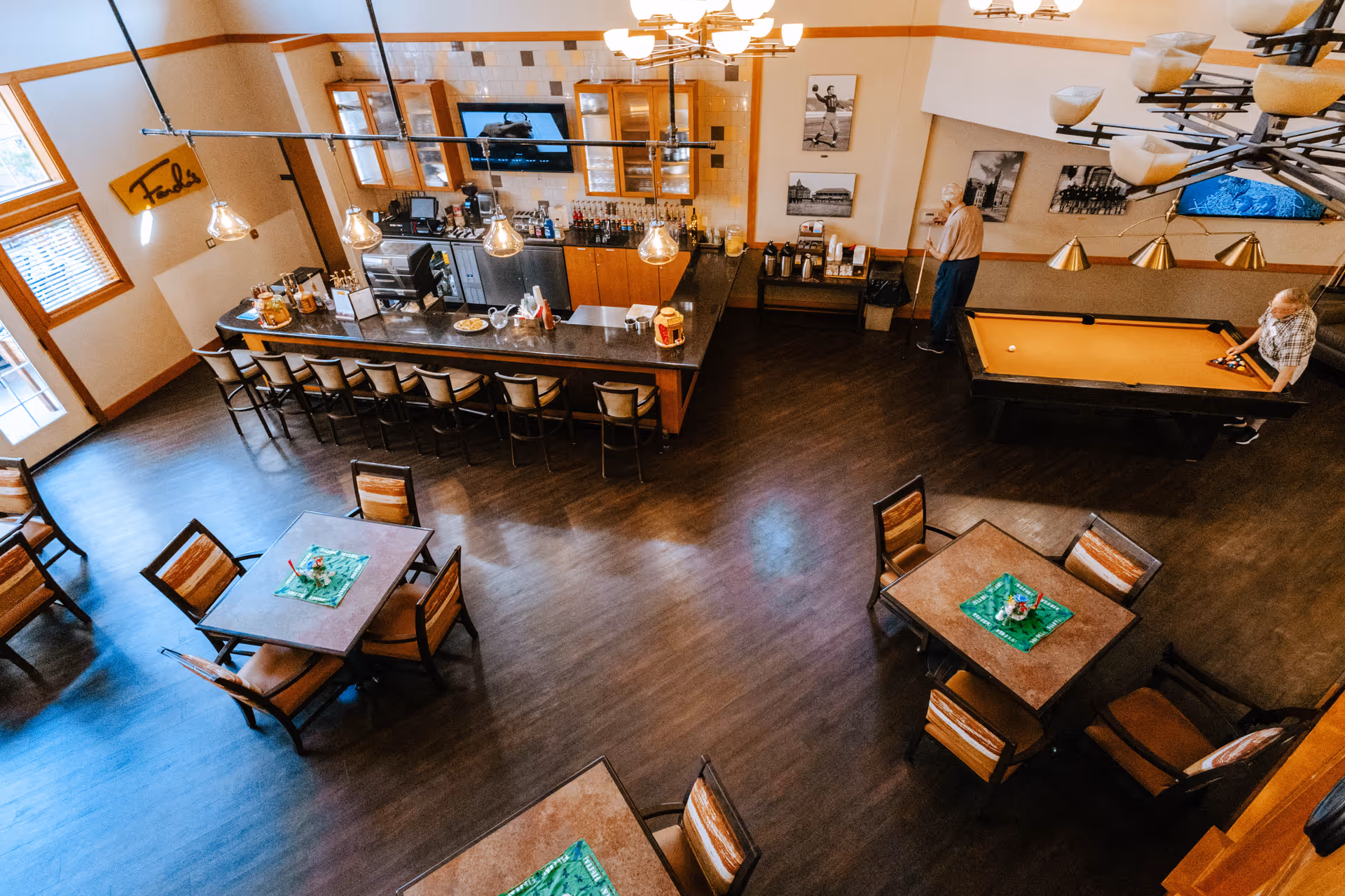 Overhead view of a senior living common area with a long bar, several dining tables, and a pool table where two residents are standing.