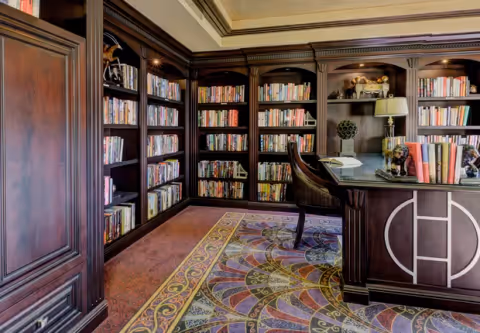A cozy library room with dark wooden bookshelves filled with books, a matching dark wooden desk with a chair, decorative items including a lamp and sculptures, and a colorful patterned carpet on the floor.