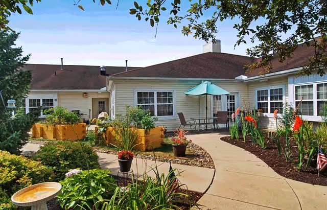 Outdoor garden area at Broadway Creek Senior Living featuring a curved concrete pathway, raised wooden garden beds with plants, potted flowers, and a patio table with chairs and a green umbrella. The building has beige siding and multiple windows under a brown roof. Trees and various plants surround the area.