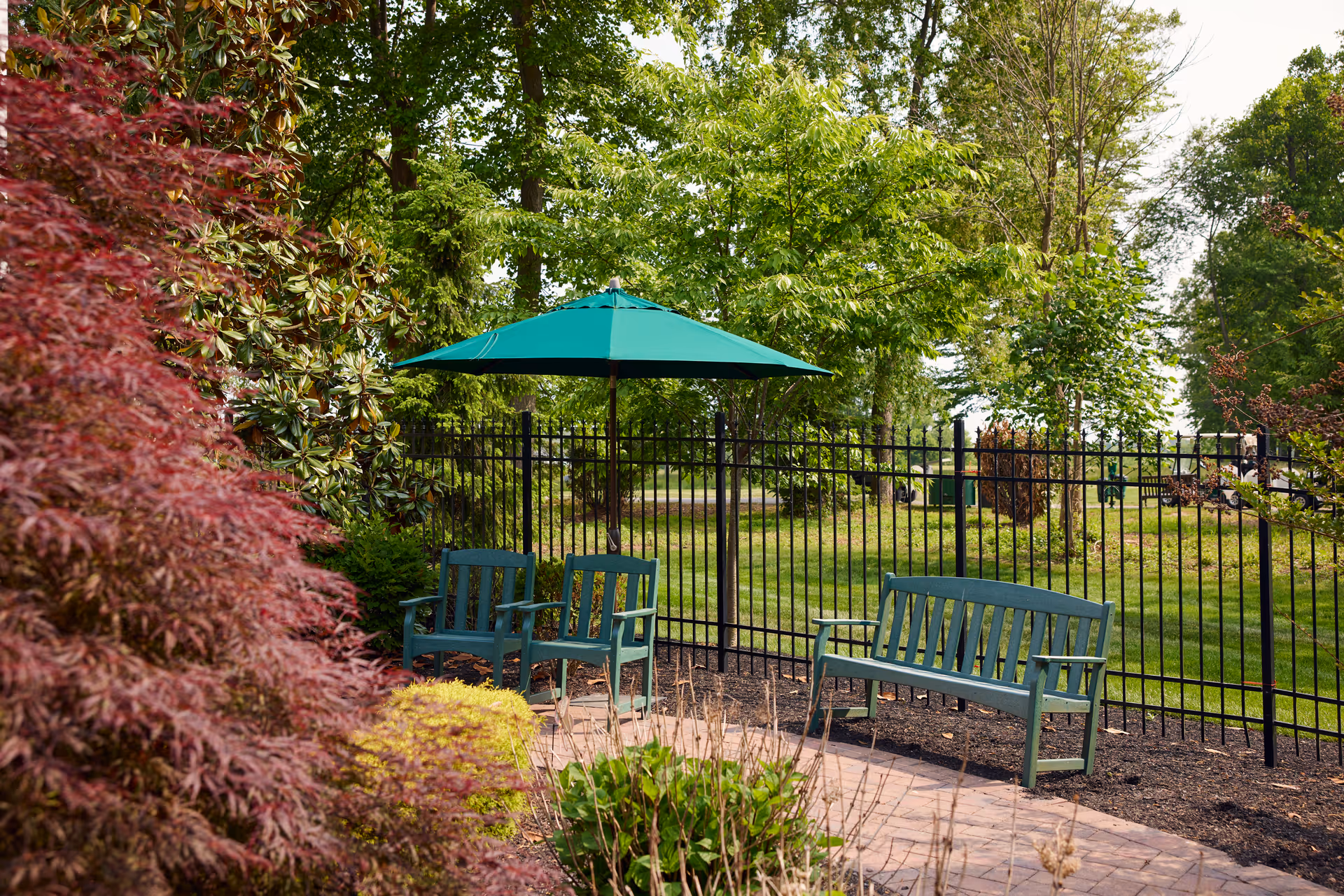 Outdoor garden area with green trees and bushes, featuring a black metal fence, two green chairs under a green patio umbrella, and a green bench on a paved surface.