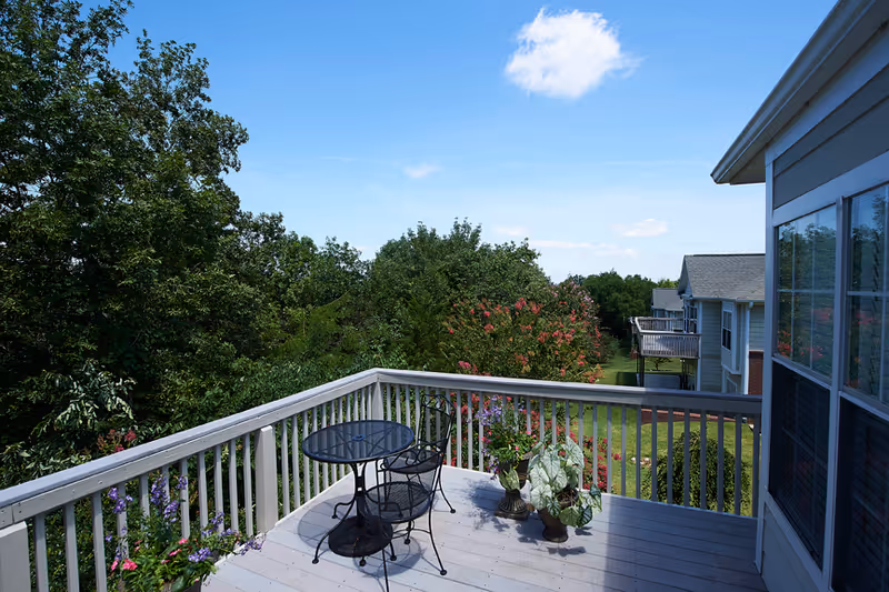 A wooden balcony with a black metal round table and two matching chairs, decorated with potted plants. The balcony overlooks a green garden area with trees and flowering bushes, and neighboring buildings with balconies are visible under a blue sky with a few clouds.