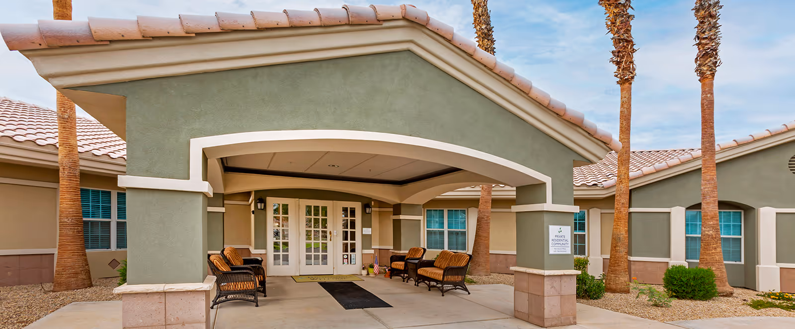 Entrance of Brookdale Camino del Sol facility featuring a covered porch with cushioned wicker chairs and a glass double door. The building has a green and beige exterior with a tiled roof and palm trees surrounding the entrance.