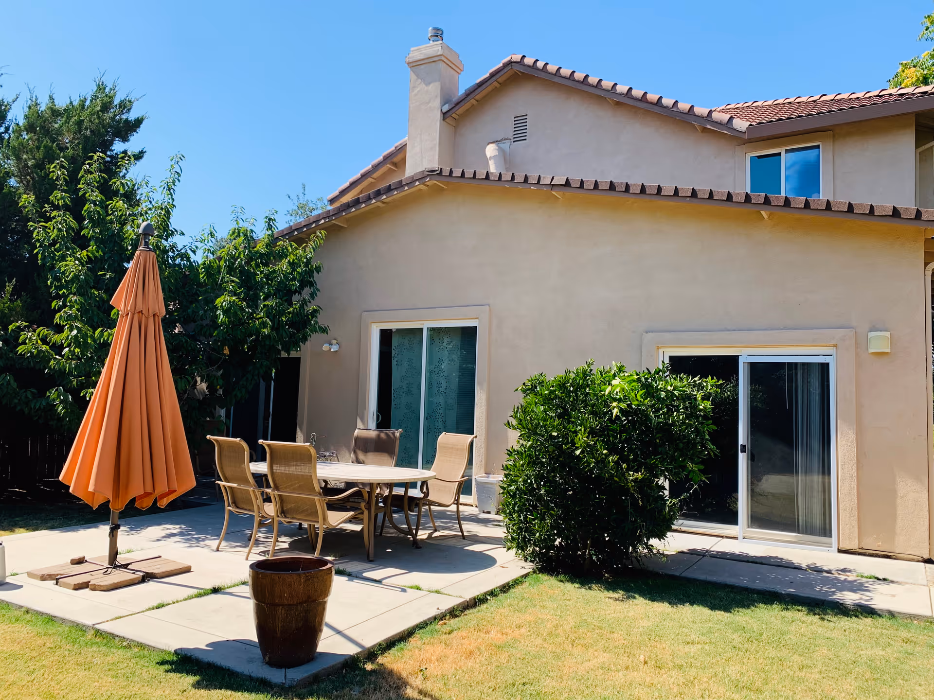 Outdoor patio area of a beige stucco building with a tiled roof, featuring a round table with six chairs and a closed orange patio umbrella. There is a green bush near a sliding glass door, and the patio is surrounded by grass and trees under a clear blue sky.
