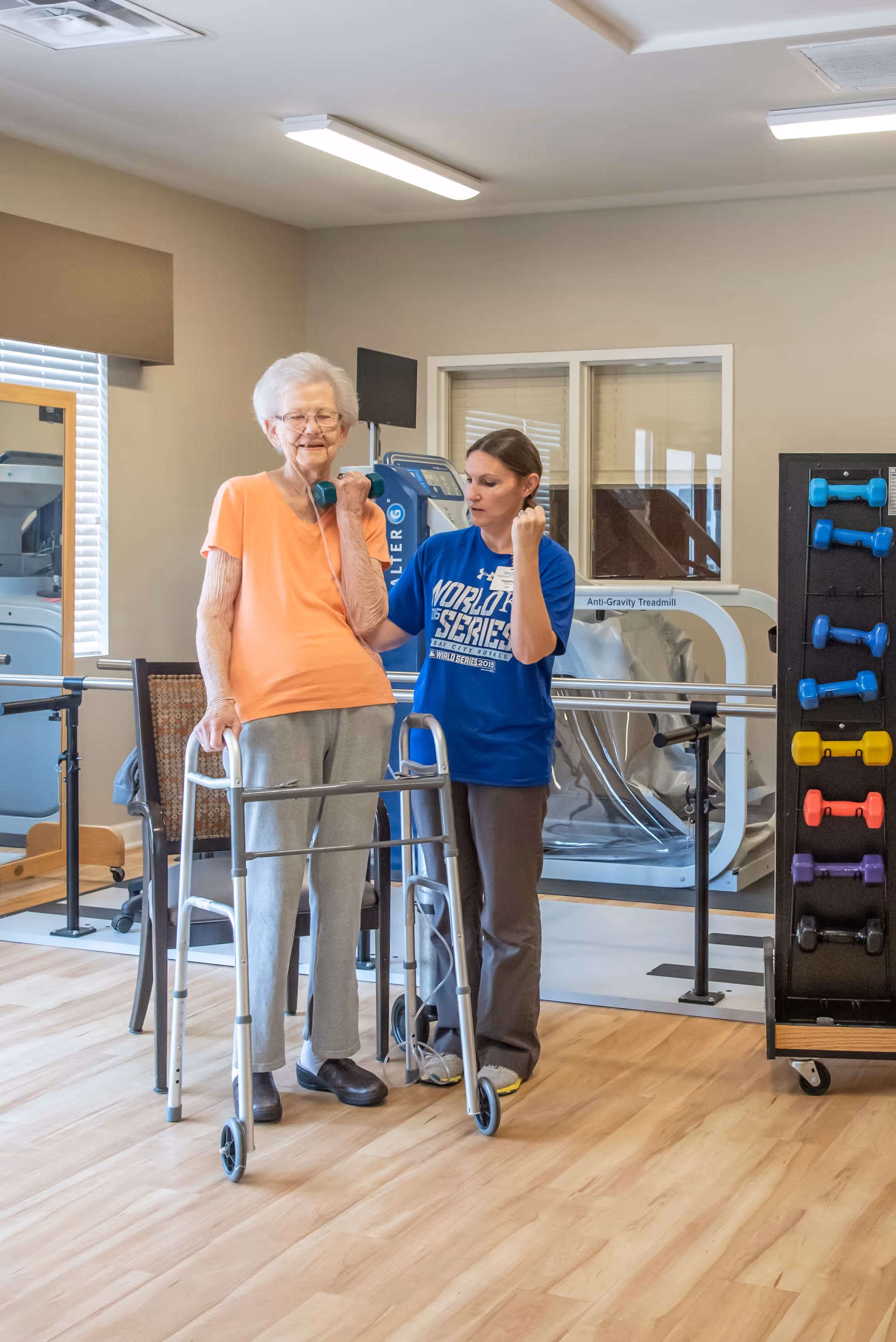 An elderly woman using a walker and holding a small dumbbell is assisted by a female physical therapist in a rehabilitation room with exercise equipment and colorful dumbbells on a rack.