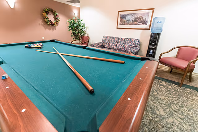 A recreational room with a green pool table in the foreground, two pool cues resting on the table, a rack of billiard balls, a floral-patterned couch, two chairs, a water cooler, a framed picture on the wall, and a decorative wreath hanging on a brown wall.