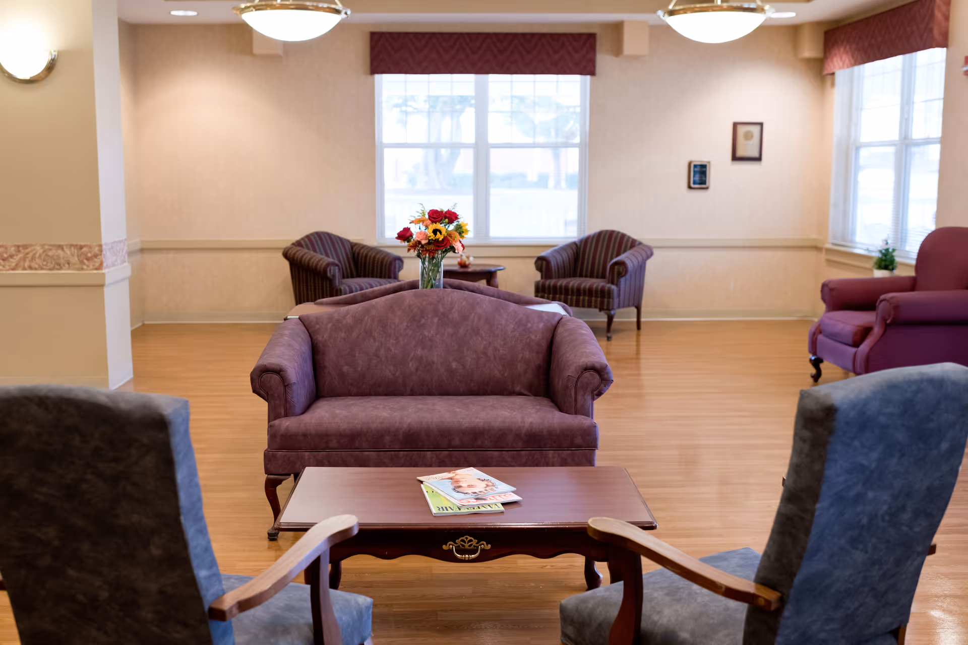 A cozy senior living facility common area with multiple upholstered chairs and sofas arranged around a wooden coffee table. The room has large windows with red valances, light-colored walls, and wood flooring. A vase with colorful flowers is placed on a table in the background.