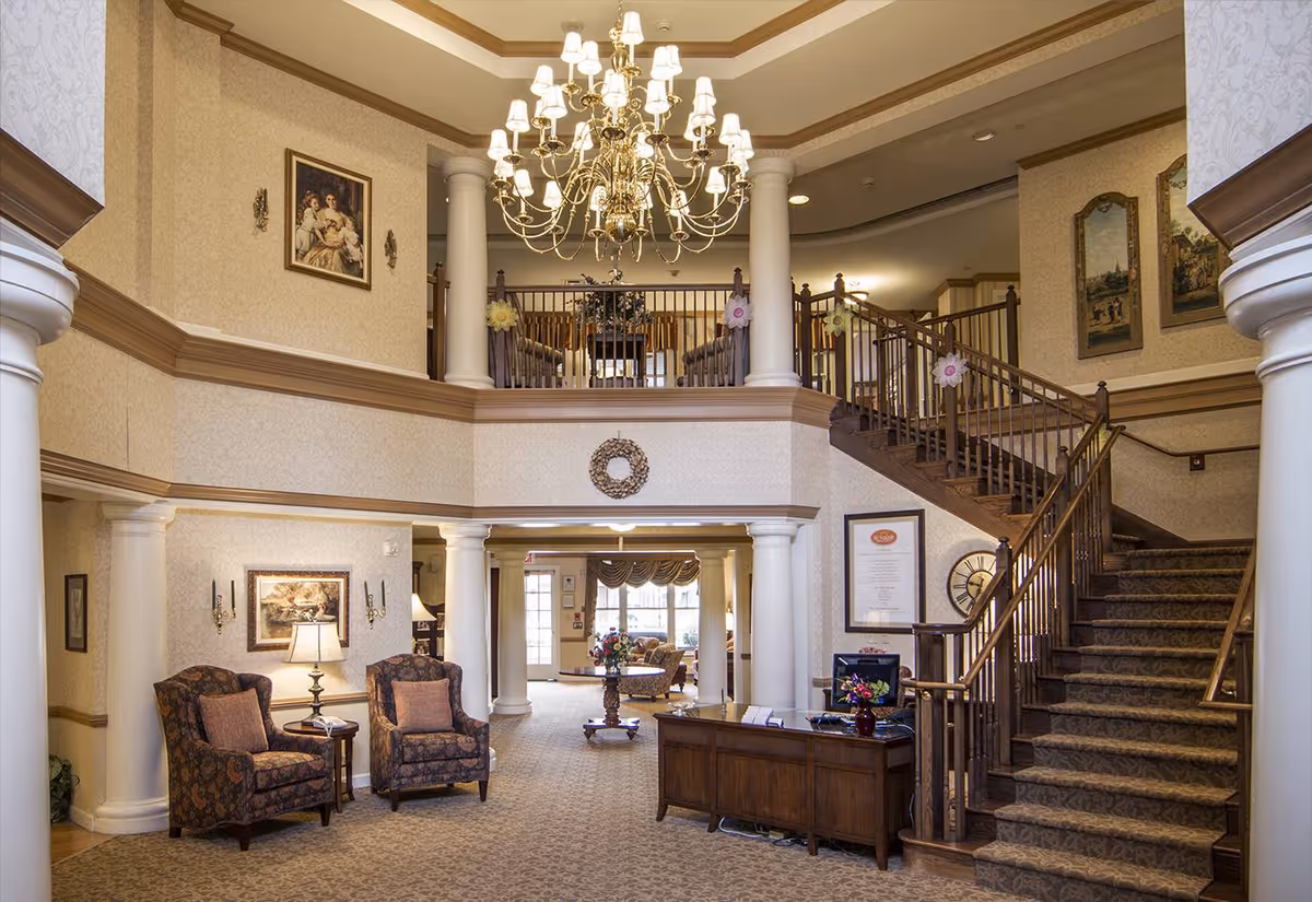 Interior view of a senior living facility lobby with a grand chandelier hanging from the ceiling, a staircase with wooden railings on the right, two upholstered armchairs with a small table and lamp on the left, and a wooden reception desk in the center. The walls are decorated with framed paintings and a wreath, and there is a seating area visible in the background.