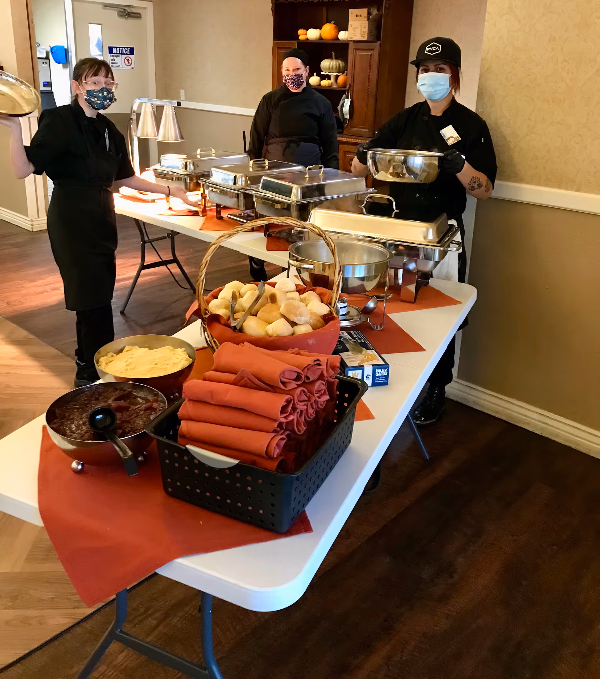 Three masked servers stand behind a buffet table with chafing dishes, a basket of rolls and folded napkins in a dining area.