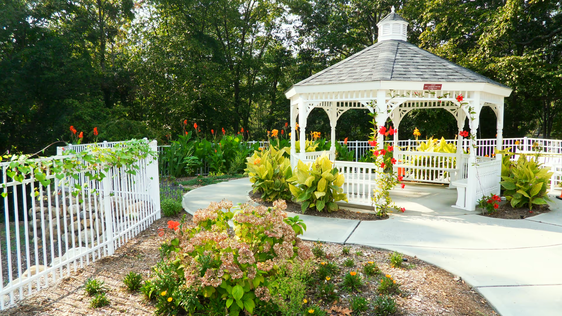 White wooden gazebo surrounded by flowering plants and a paved walkway in a landscaped garden.