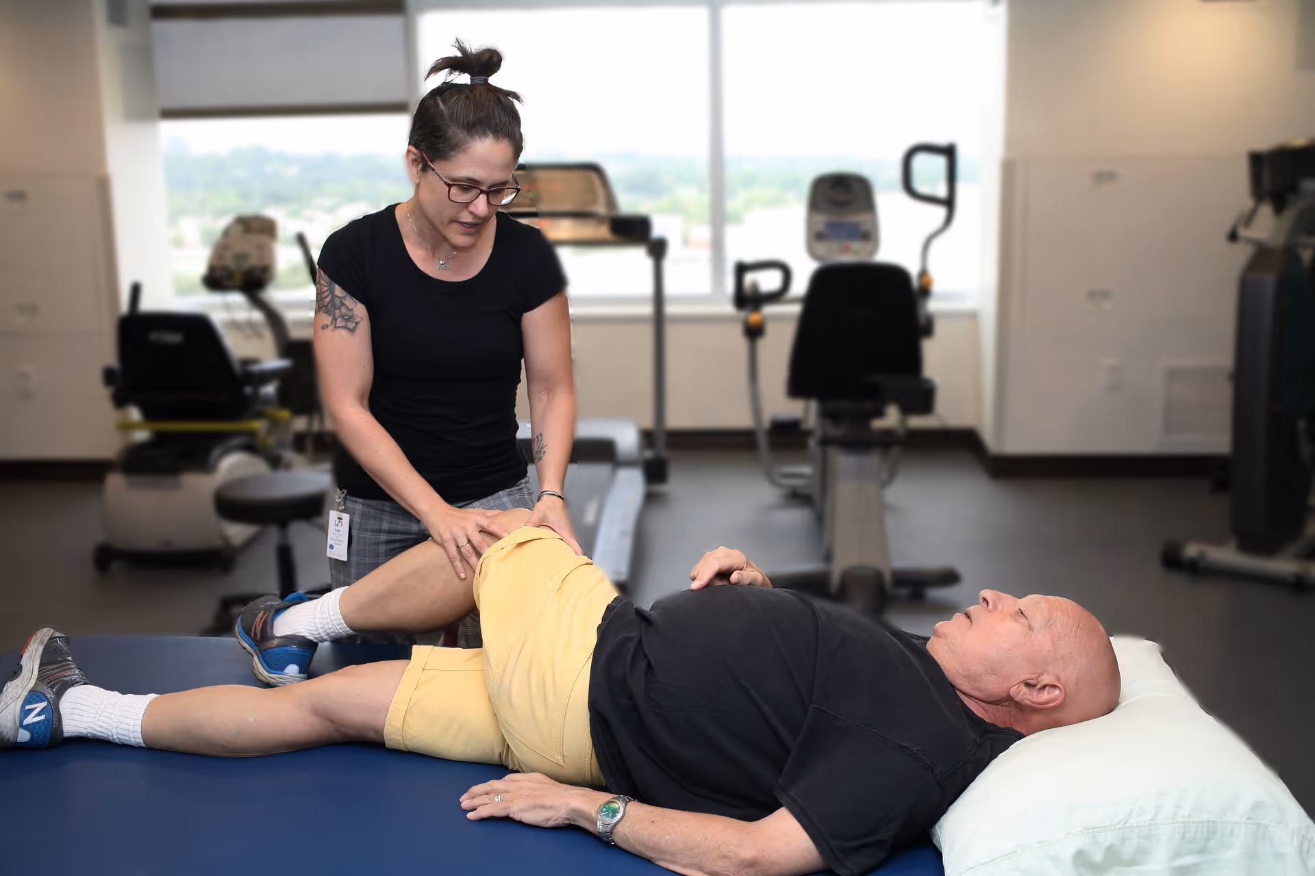 A woman wearing glasses and a black shirt assists an elderly man lying on a padded table in a fitness or rehabilitation room. The man is lying on his side with a pillow under his head, wearing a black shirt and yellow shorts. Exercise equipment is visible in the background near large windows.