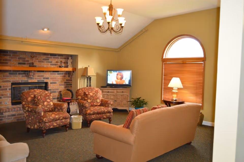 A cozy living room with beige walls and carpeted floor featuring a brick fireplace, two patterned armchairs, a beige sofa, a wooden cabinet with a TV on top, a side table with a lamp, and a large arched window with wooden blinds partially closed.