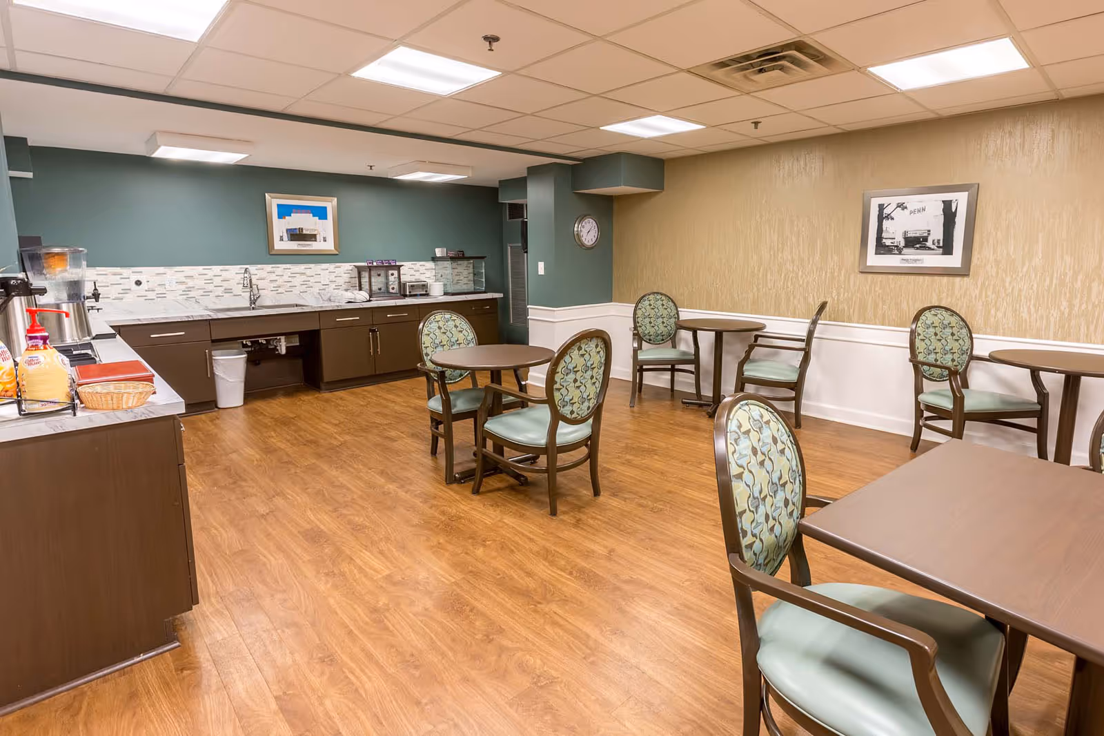 A small dining area with several round tables and chairs with patterned backs and green seats. The room has wood flooring, a green accent wall with a framed picture, and a beige wall with another framed picture. There is a countertop with a sink, a water dispenser, and some condiments on the left side of the room.