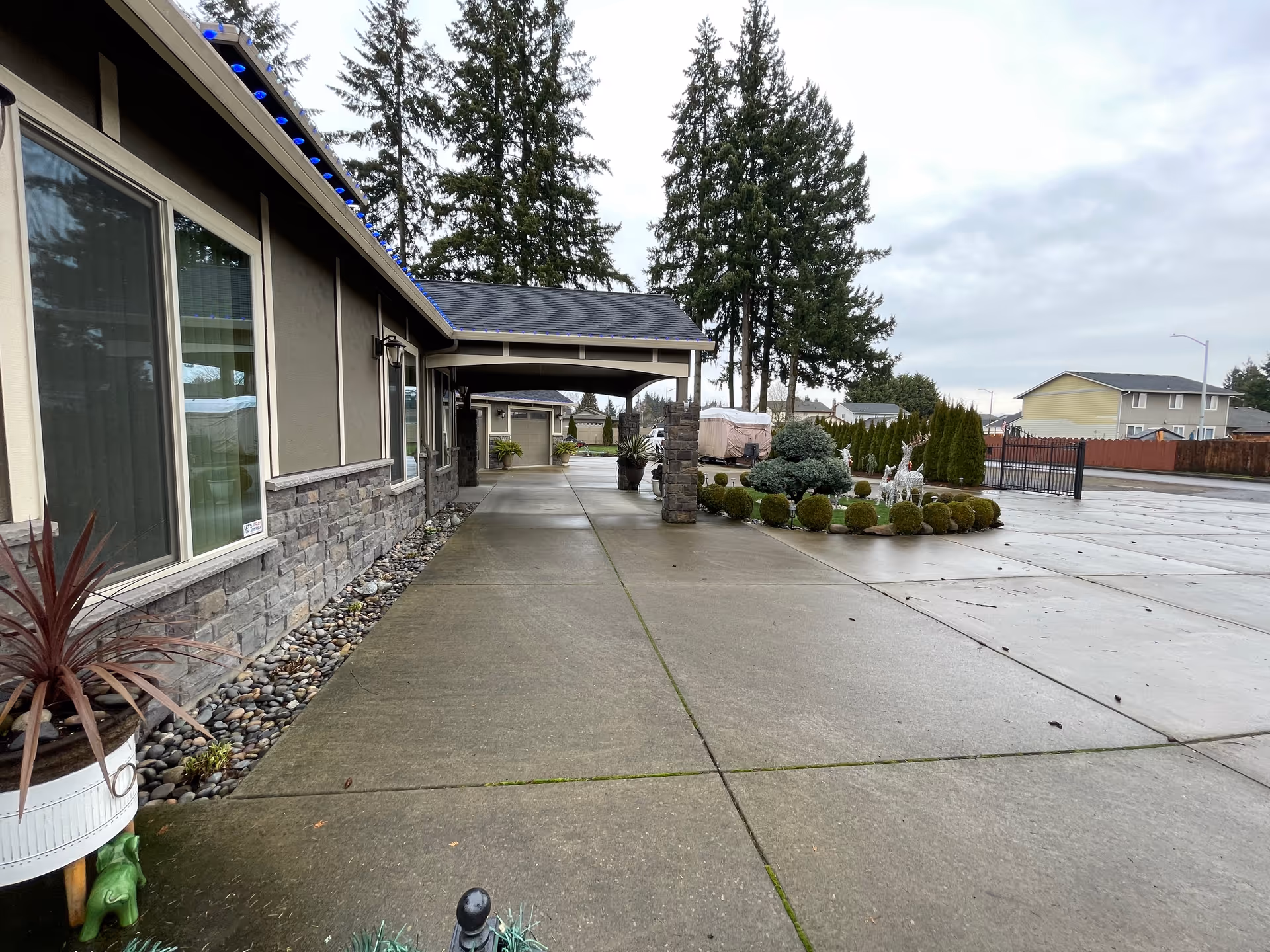Exterior view of Nataly’s Adult Family Home showing a paved walkway alongside the building with stone and beige siding. There are decorative plants and bushes near the walkway and a gated driveway in the background under a cloudy sky.