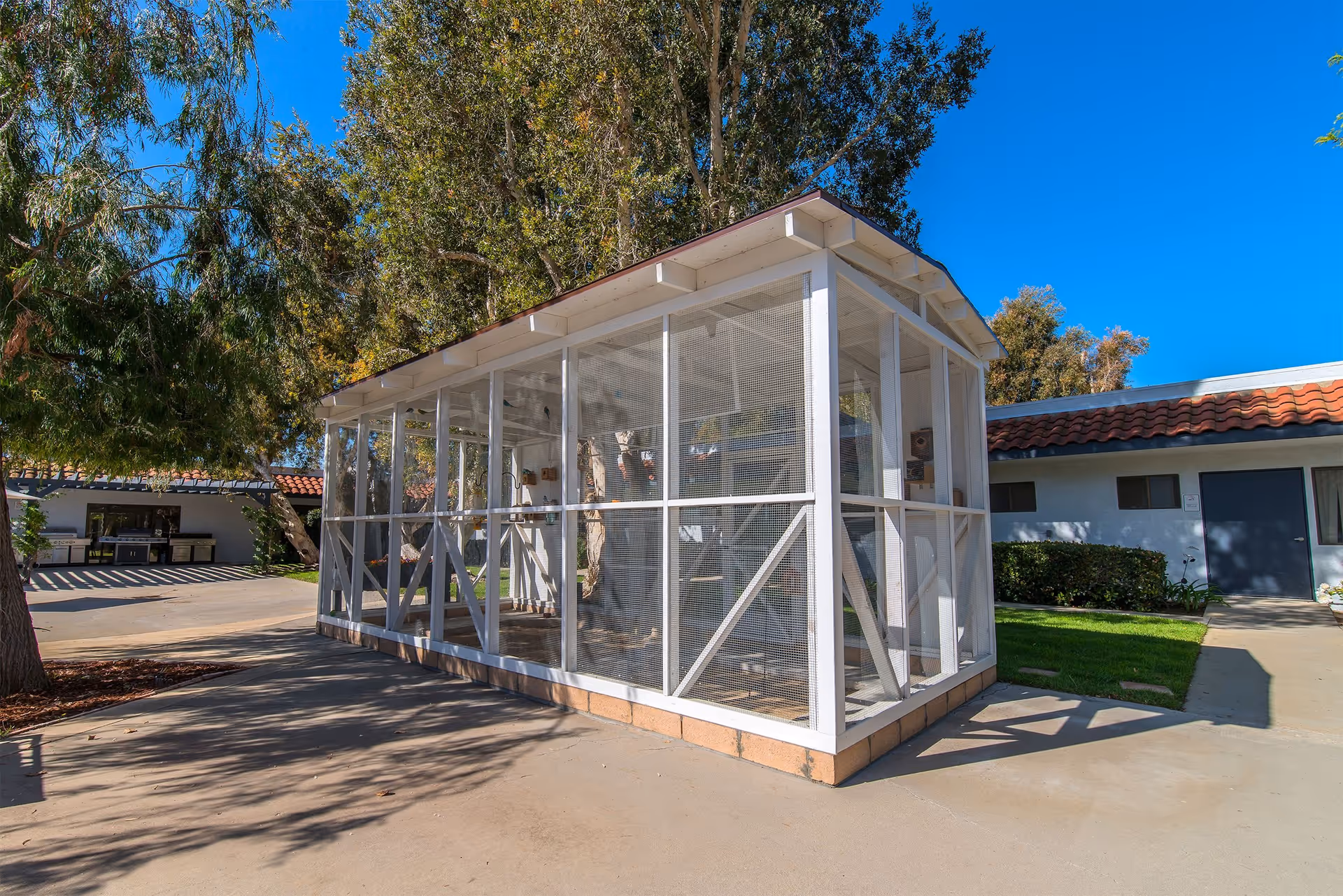 Outdoor view of a white screened enclosure structure with a pitched roof, surrounded by trees and adjacent to a building with a red tile roof under a clear blue sky.