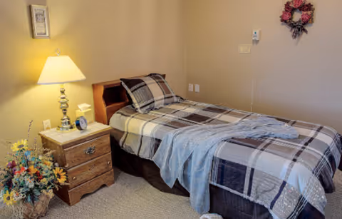 A simple single-occupancy bedroom with a twin bed dressed in plaid bedding, a wooden nightstand with a lamp, and a floral arrangement.