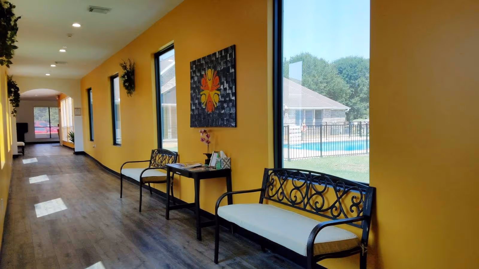 Sunlit interior hallway with yellow walls, decorative benches and windows overlooking a pool.