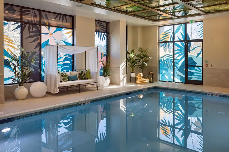 Indoor swimming pool area with large windows decorated with colorful leaf and flower patterns. There is a white canopy daybed with green and blue pillows, potted plants, a large golden duck decoration, and round white lights around the poolside.