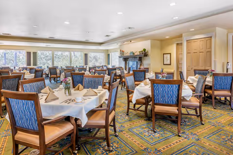 A spacious dining room with multiple tables covered in white tablecloths, each set with folded napkins, cups, and small flower arrangements. The room has large windows letting in natural light, patterned carpet, and wooden chairs with blue upholstered backs and tan seats.