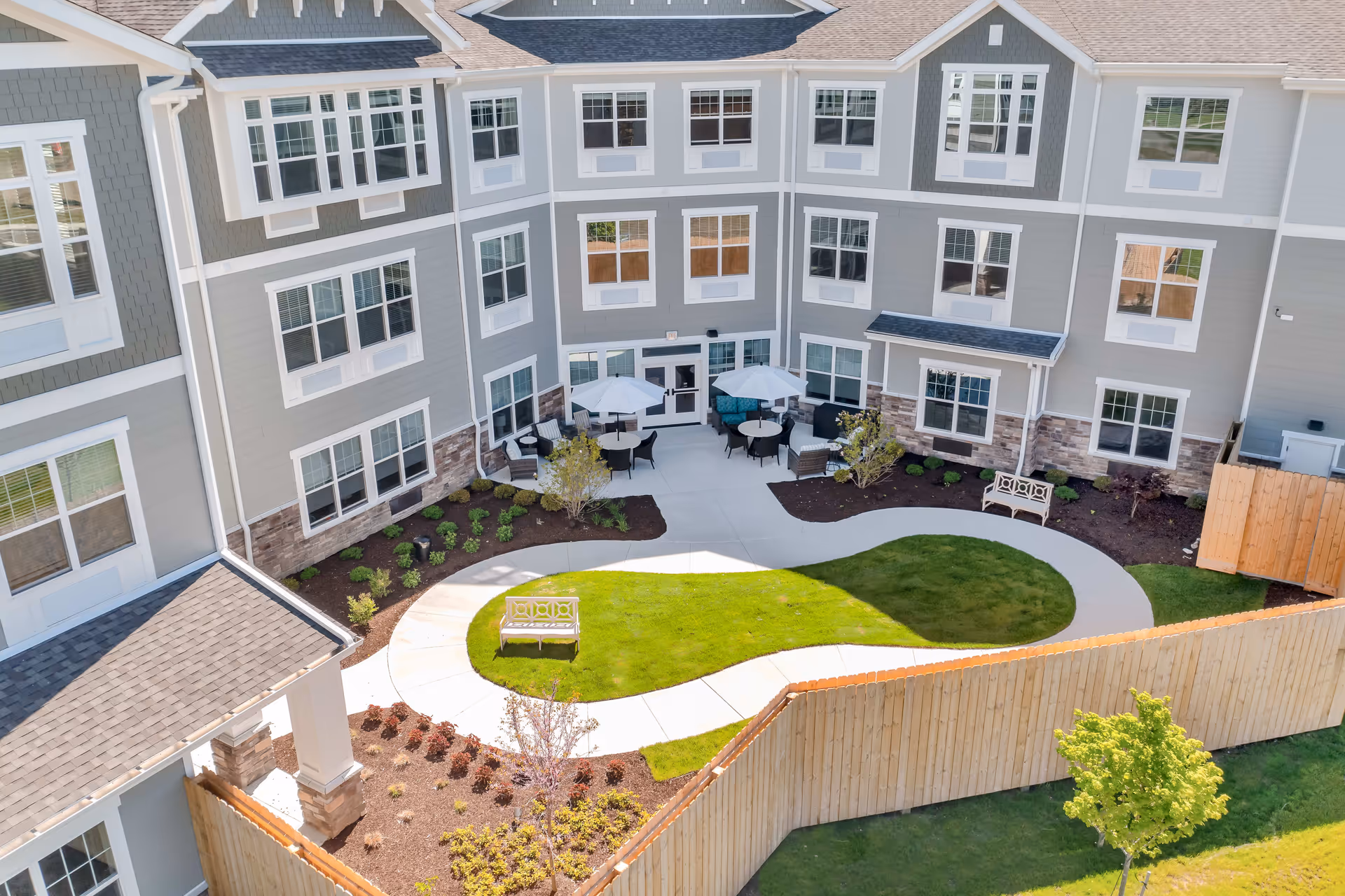 A courtyard area of a senior living facility with a curved concrete walkway surrounding a grassy lawn. There are benches along the walkway and several tables with umbrellas and chairs near the building. The building has multiple windows and gray siding with white trim. A wooden fence encloses the courtyard.