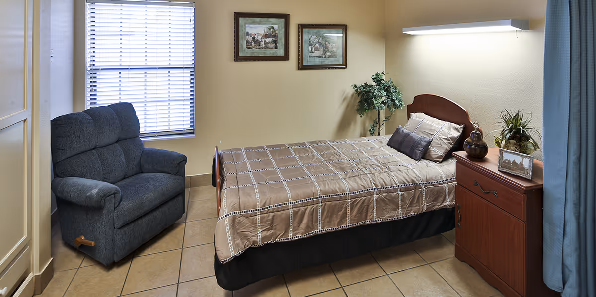 Small furnished bedroom with a single bed, bedside cabinet, recliner chair, window with blinds and wall art.