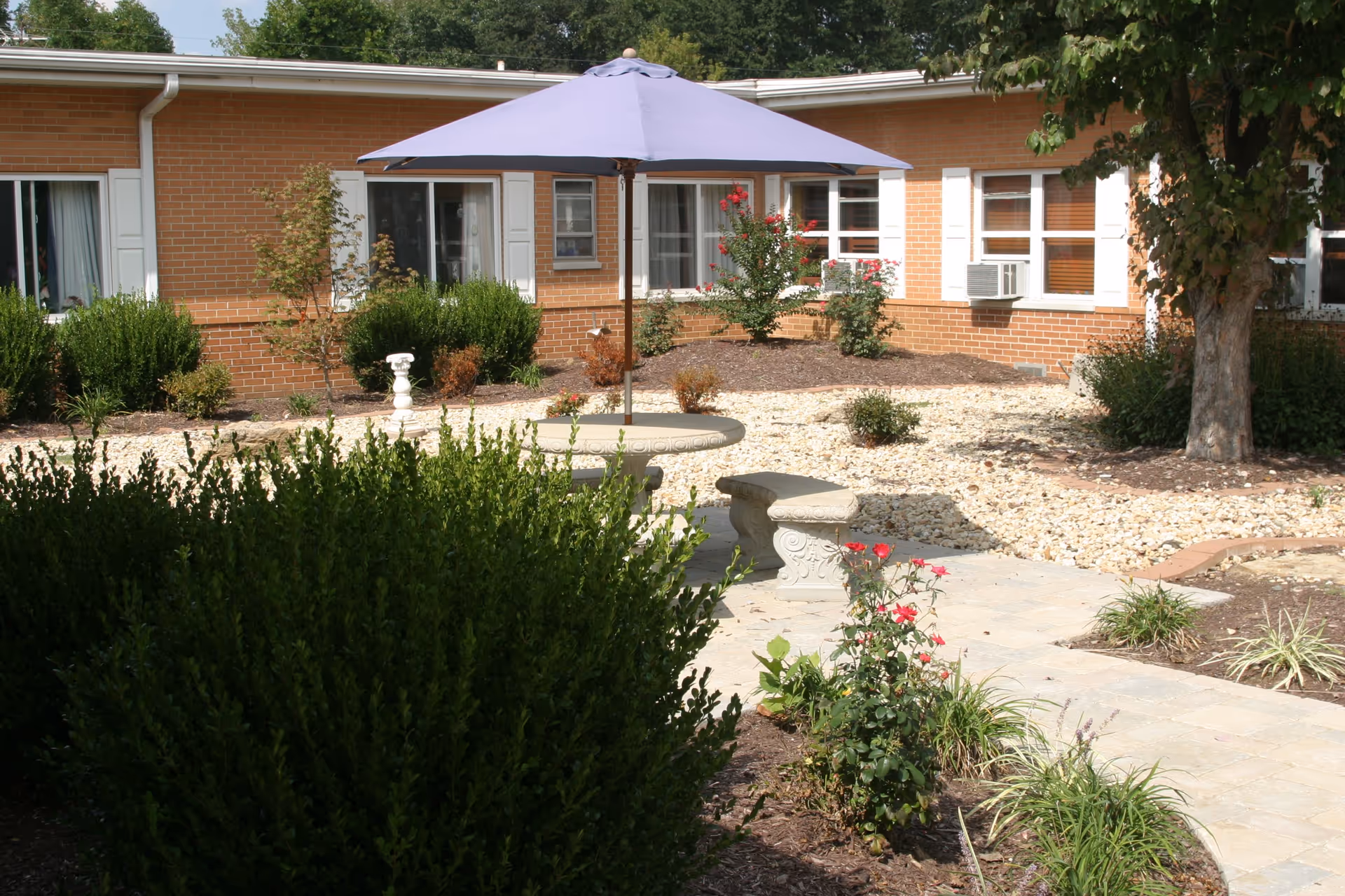 Outdoor courtyard area of Sycamore Village Assisted Living featuring a round stone table with a large purple umbrella and two stone benches. The courtyard is landscaped with bushes, flowering plants, and a tree, surrounded by a brick building with multiple windows.