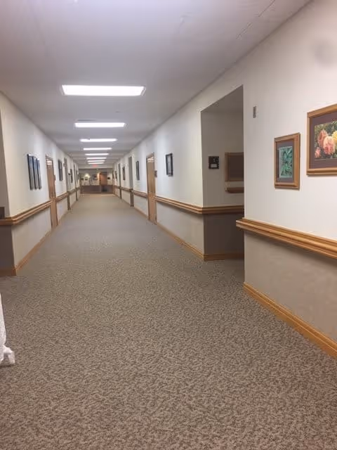 A long, carpeted hallway in a senior living facility with beige walls, wooden trim, and framed artwork hanging on the walls. The ceiling has recessed fluorescent lighting, and there are handrails along both sides of the hallway.