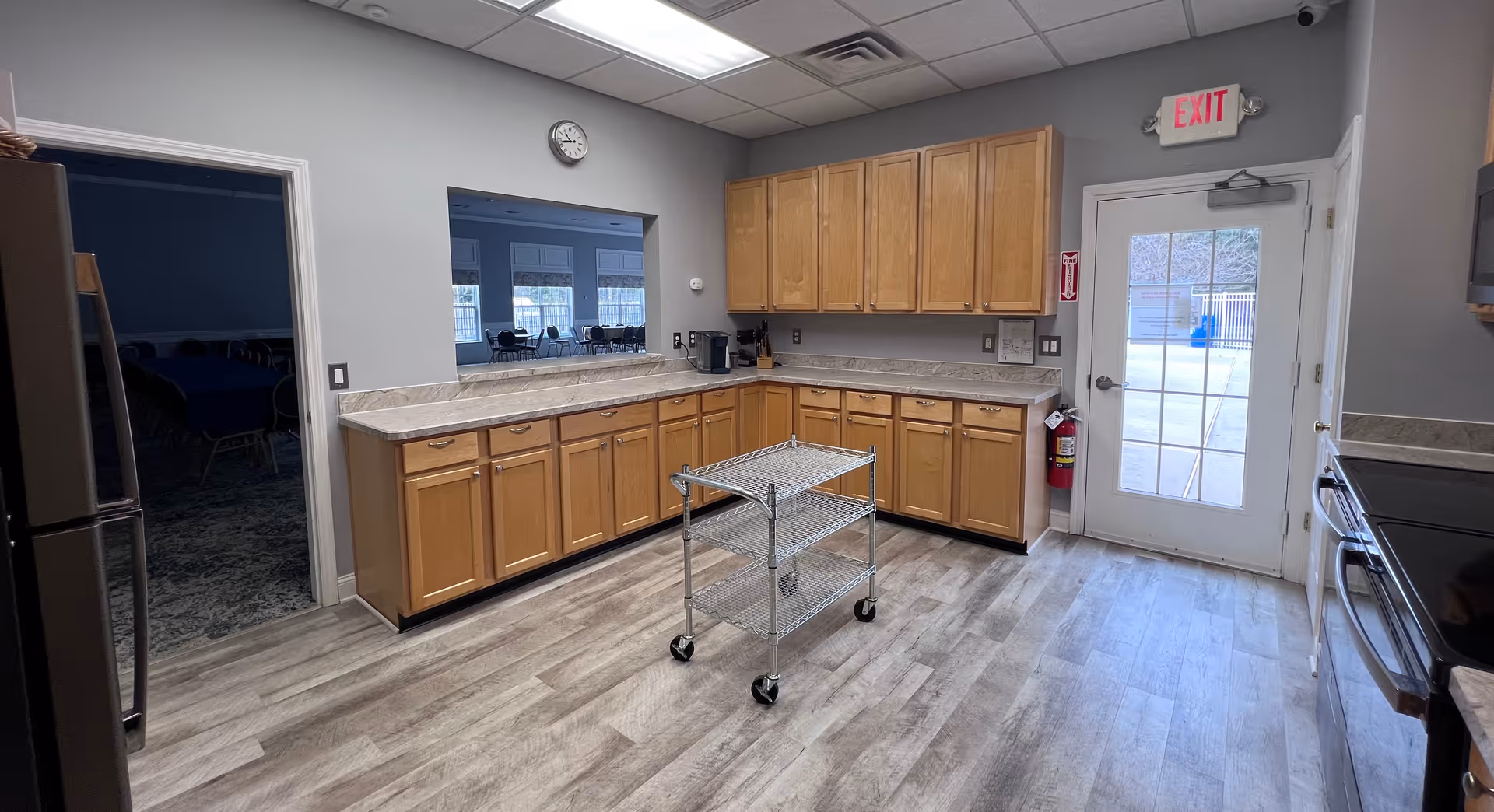 A clean kitchen area with light wood cabinets, marble countertops, and a small metal rolling cart in the center. There is a door with a window leading outside, a clock on the wall, and an open pass-through window to an adjacent room with tables and chairs. The floor is light wood laminate, and the walls are painted light gray.