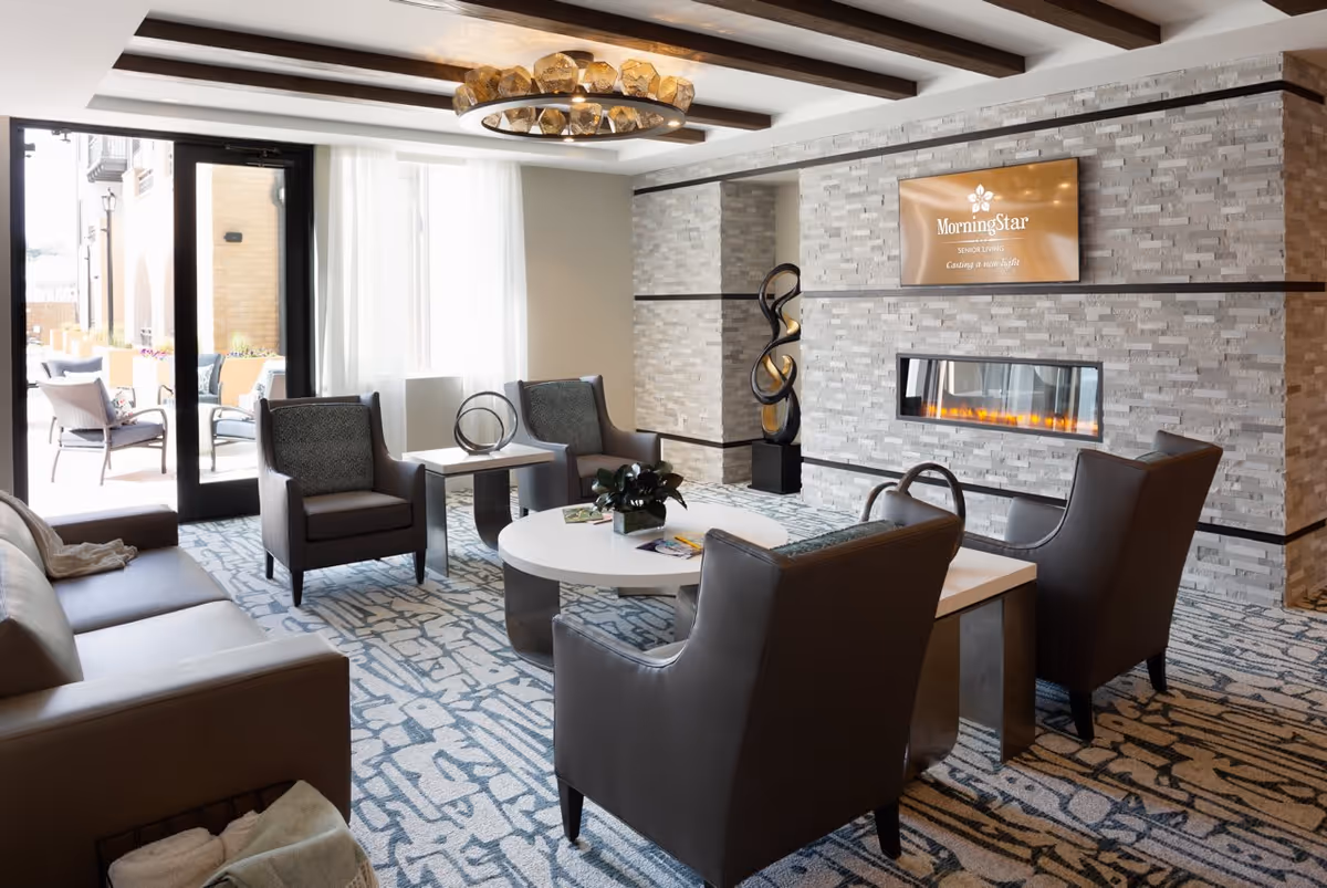 A modern senior living facility lounge area with a stone accent wall featuring a built-in electric fireplace and a mounted screen displaying the MorningStar Senior Living logo. The room has a patterned carpet, leather armchairs, a sofa, a round coffee table with a plant, and a large window with sheer curtains letting in natural light. There is also a decorative sculpture and an outdoor seating area visible through glass doors.